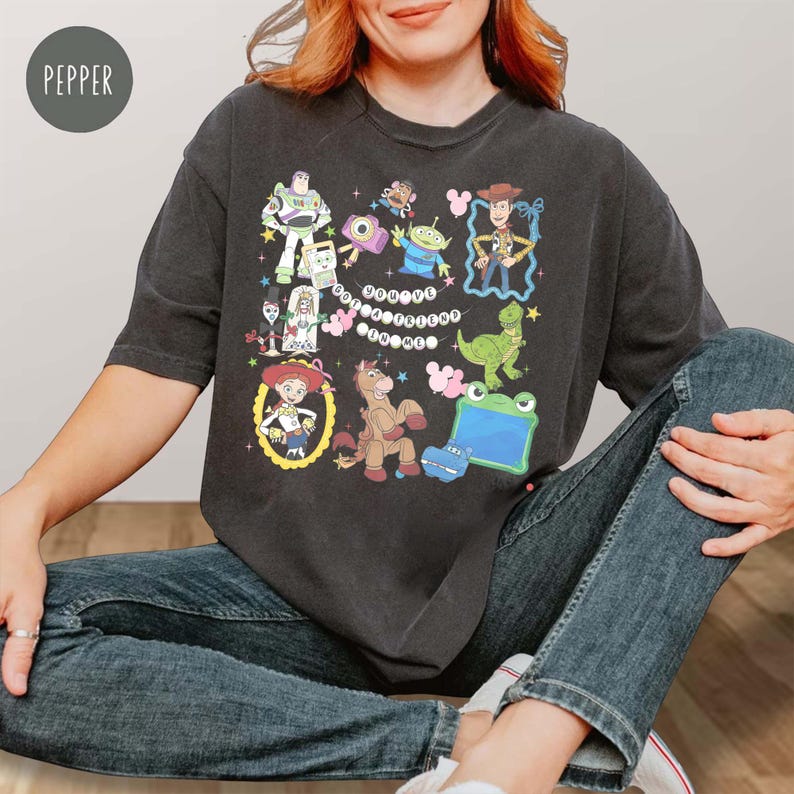 May include: A charcoal gray short-sleeve t-shirt featuring a colorful graphic print of various Toy Story characters including Woody, Buzz Lightyear, Jessie, and Rex, with the text "YOU'VE GOT A FRIEND IN ME" centered among the character illustrations.