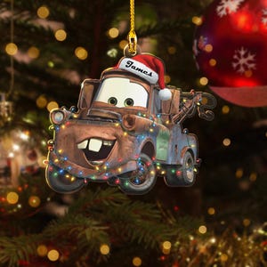 May include: A Christmas tree decoration depicting the character Mater from the film Cars. The brown tow truck wears a Santa hat with the name "James" and is adorned with colourful Christmas lights. A red bauble is in the background.