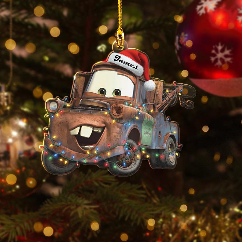 May include: A Christmas tree decoration depicting the character Mater from the film Cars. The brown tow truck wears a Santa hat with the name "James" and is adorned with colourful Christmas lights. A red bauble is in the background.