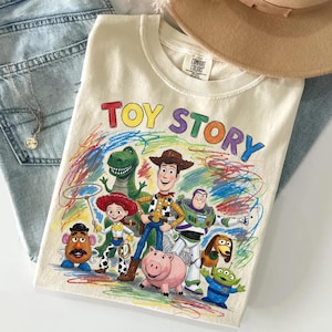 Peut inclure: T-shirt cr&egrave;me avec l'inscription "TOY STORY" en lettres color&eacute;es au-dessus d'un dessin des personnages du film d'animation. Le motif comprend Woody, Buzz l'&Eacute;clair et d'autres personnages. Le t-shirt est pli&eacute; sur un jean bleu.