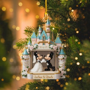May include: A decorative ornament featuring Mickey and Minnie Mouse in wedding attire, set against a castle backdrop. The ornament is adorned with the word "ENGAGED" and the names "Luciana & David". The castle has pink and blue accents.