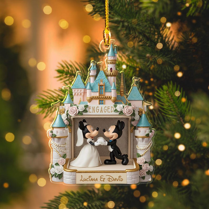 May include: A decorative ornament featuring Mickey and Minnie Mouse in wedding attire, set against a castle backdrop. The ornament is adorned with the word "ENGAGED" and the names "Luciana & David". The castle has pink and blue accents.