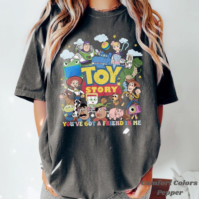 Puede incluir: Una camiseta de manga corta gris carb&oacute;n con un estampado gr&aacute;fico colorido de personajes de Toy Story, incluidos Buzz Lightyear, Woody y Jessie. El texto dice TOY STORY y YOU'VE GOT A FRIEND IN ME, rodeado de estrellas y nubes.