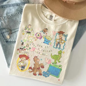 May include: A cream-colored t-shirt featuring a graphic print of various Toy Story characters including Woody, Buzz Lightyear, Jessie, Rex, and Bullseye. The text "YOU'VE GOT A FRIEND IN ME" is printed in the center, surrounded by small stars and balloons.