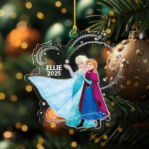 May include: A clear acrylic Christmas ornament featuring Elsa and Anna from Frozen, with the name "Ellie" and the year "2025" printed on it. The ornament is shaped like a snowflake and is hanging from a gold string.