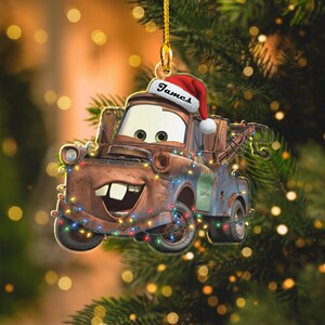 May include: A Christmas tree decoration of the character Mater from the film Cars, wearing a Santa hat and fairy lights. The name "James" is written on the hat. The ornament is brown, blue, and red.