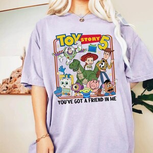 May include: A light purple t-shirt featuring a colorful graphic of characters from the movie Toy Story 5. The graphic includes Buzz Lightyear, Woody, and other characters. The text "TOY STORY 5" and "YOU'VE GOT A FRIEND IN ME" are also displayed.