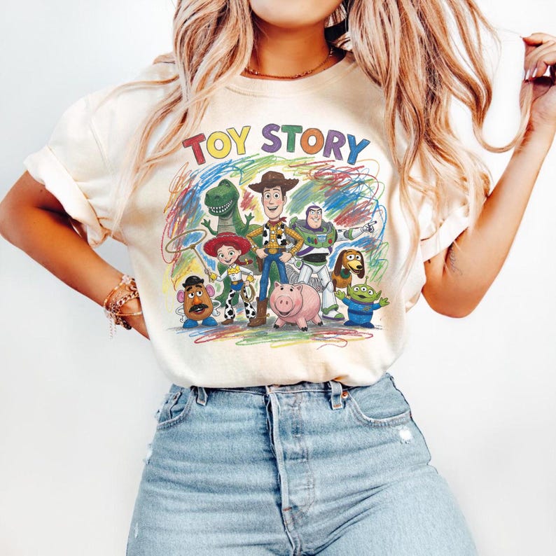 Peut inclure: T-shirt cr&egrave;me avec un graphisme color&eacute; des personnages de Toy Story, dont Woody, Buzz l'&Eacute;clair et Rex. Les mots "TOY STORY" sont imprim&eacute;s au-dessus des personnages dans une police arc-en-ciel. Le design est entour&eacute; de gribouillis color&eacute;s.