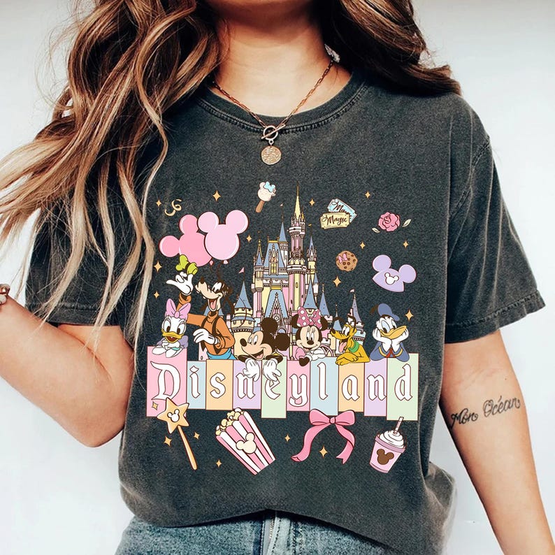 Puede incluir: Camiseta gris oscuro con un dise&ntilde;o colorido de Disneyland. El dise&ntilde;o incluye a Mickey y Minnie Mouse, el Pato Donald, Goofy y Daisy, un castillo, globos y la palabra "Disneyland".