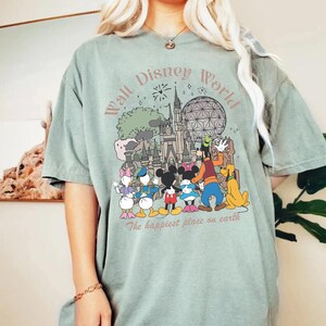May include: Sage green t-shirt featuring a colorful graphic of Disney characters and landmarks, including Mickey Mouse, Donald Duck, and Cinderella's castle. The text "Walt Disney World" and "The happiest place on earth" are also displayed.