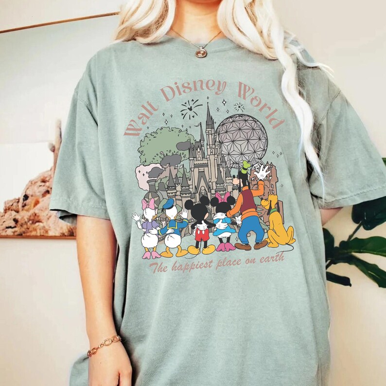 May include: Sage green t-shirt featuring a colorful graphic of Disney characters and landmarks, including Mickey Mouse, Donald Duck, and Cinderella's castle. The text "Walt Disney World" and "The happiest place on earth" are also displayed.