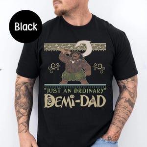May include: Black t-shirt featuring a cartoon illustration of a character holding a hook. The text reads "JUST AN ORDINARY DEMI-DAD" in a stylized font. The design includes decorative elements.