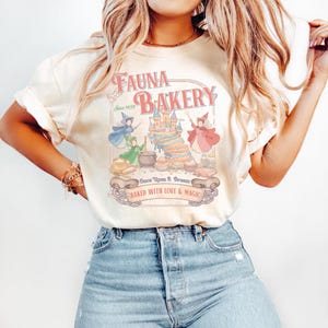 May include: Cream-colored t-shirt featuring the text "Fauna Bakery" with a vintage-style design. The graphic includes illustrations of fairies, a castle-shaped cake, and the phrase "Baked with Love & Magic."