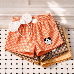 May include: A pair of orange and white gingham shorts featuring a small white ghost graphic with Mickey Mouse ears, paired with a white headband adorned with white floral-shaped Mickey Mouse ears and a white bow, layered over brown and yellow shorts.