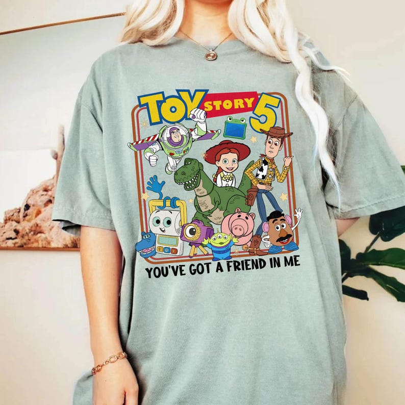 May include: A light green t-shirt featuring a colorful graphic of characters from Toy Story 5. The design includes Buzz Lightyear, Woody, and other characters, with the text "YOU'VE GOT A FRIEND IN ME".