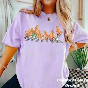 Może przedstawiać: Jasnofioletowa koszulka z grafiką postaci z kresk&oacute;wek przechodzących przez przejście dla pieszych, z pomarańczowym pachołkiem na głowie. U dołu nadruk "Comfort Colors Orchid". Koszulka ma luźny kr&oacute;j.