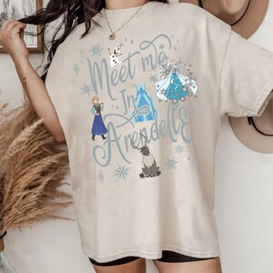 Puede incluir: Camiseta beige con el texto "Meet me in Arendelle" en escritura azul claro. La camiseta presenta ilustraciones de personajes de la pel&iacute;cula Frozen, incluyendo Elsa, Anna, Olaf y Sven, junto con copos de nieve. La camiseta tiene un ajuste casual y de gran tama&ntilde;o.