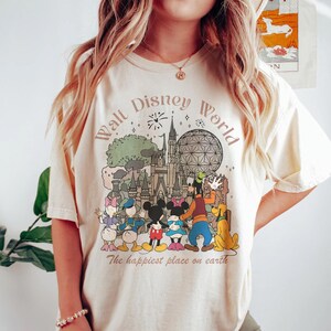 May include: Cream-colored t-shirt featuring a graphic of Disney characters and landmarks, including Mickey and Minnie Mouse, Goofy, and Pluto. The text "Walt Disney World" and "The happiest place on earth" are also displayed.
