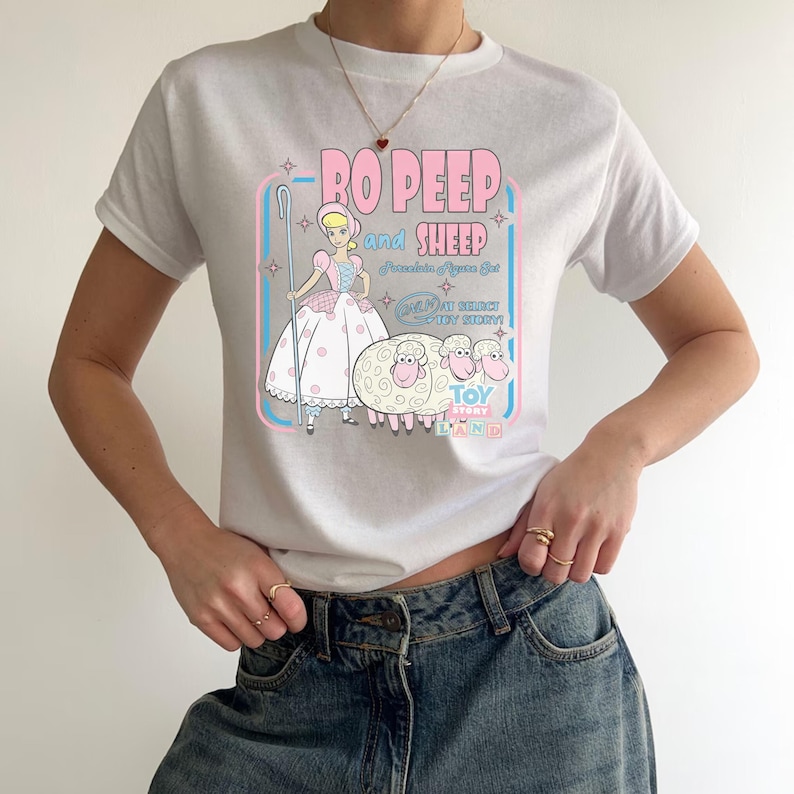以下が含まれることがあります： トイ・ストーリーのボー・ピープと羊のグラフィックがプリントされた白いTシャツ。デザインには、ピンクと青で「Bo Peep and Sheep」の文字と、ボー・ピープと3匹の羊のイラストが描かれています。Tシャツは半袖です。