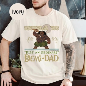 May include: Ivory-colored t-shirt featuring a cartoon illustration of a muscular character holding a hook. The shirt has the text "JUST AN ORDINARY DEMI-DAD" in a stylized font. The shirt is a casual wear item.