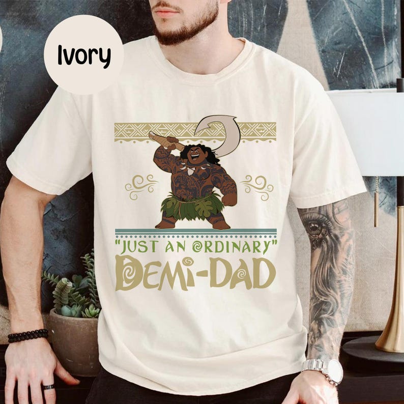 May include: Ivory-colored t-shirt featuring a cartoon illustration of a muscular character holding a hook. The shirt has the text "JUST AN ORDINARY DEMI-DAD" in a stylized font. The shirt is a casual wear item.