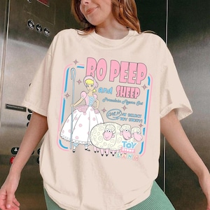 以下が含まれることがあります： クリーム色のTシャツ。トイ・ストーリーのボー・ピープと羊のグラフィックが特徴です。「Bo Peep and Sheep」の文字がピンク色で、青と白のテキストとイラストが追加されています。