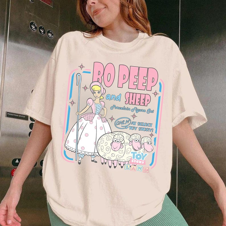 以下が含まれることがあります： クリーム色のTシャツ。トイ・ストーリーのボー・ピープと羊のグラフィックが特徴です。「Bo Peep and Sheep」の文字がピンク色で、青と白のテキストとイラストが追加されています。