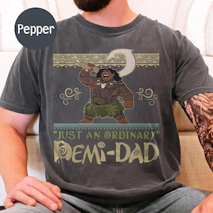 May include: Dark gray t-shirt featuring a cartoon illustration of a Polynesian character holding a hook. The shirt has the text "JUST AN ORDINARY DEMI-DAD" in a stylized font. The design includes decorative tribal patterns.