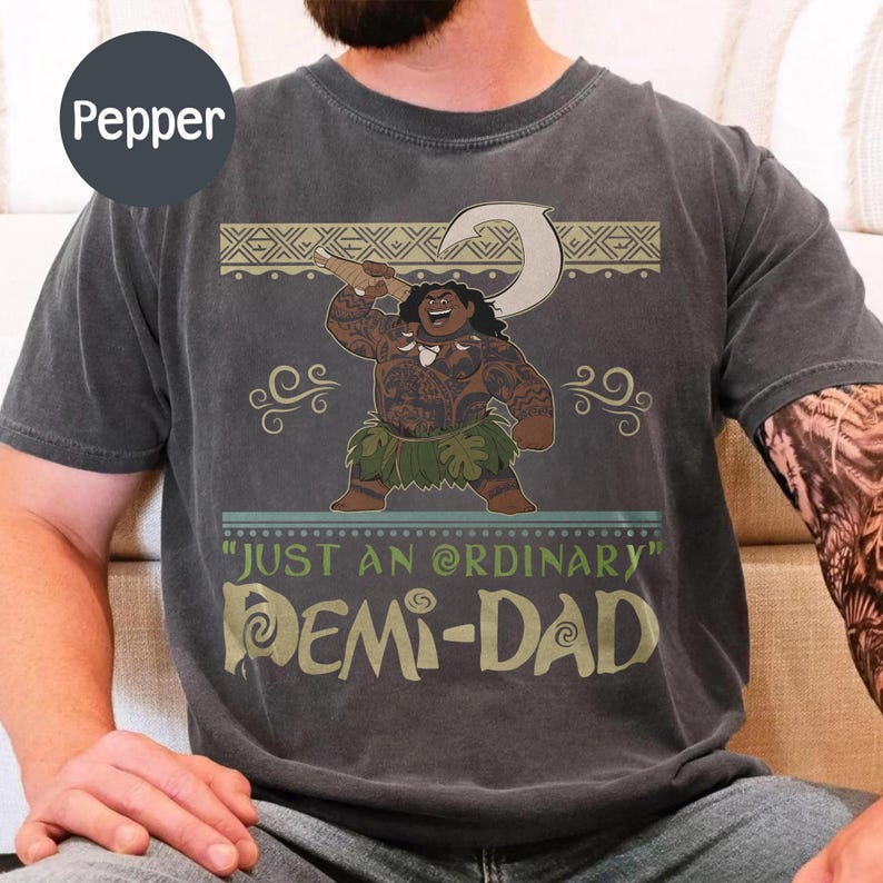 May include: Dark gray t-shirt featuring a cartoon illustration of a Polynesian character holding a hook. The shirt has the text "JUST AN ORDINARY DEMI-DAD" in a stylized font. The design includes decorative tribal patterns.