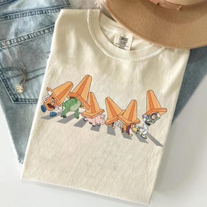 Może przedstawiać: Kremowa koszulka z zabawną grafiką postaci z kresk&oacute;wek, z kt&oacute;rych każda balansuje na głowie pomarańczowym pachołkiem drogowym. Koszulka jest złożona, z brązowym kapeluszem i niebieskimi dżinsami w tle.