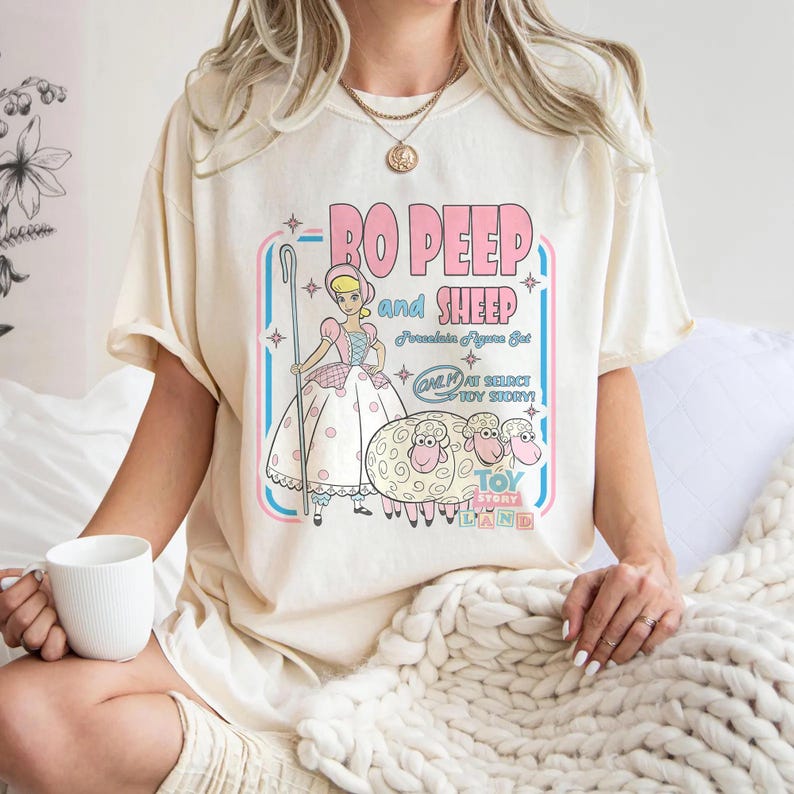 以下が含まれることがあります： トイ・ストーリーのボー・ピープと羊のグラフィックが描かれたクリーム色のTシャツ。「Bo Peep and Sheep」の文字がピンクと青でデザインされ、追加のテキストとイラストも含まれています。