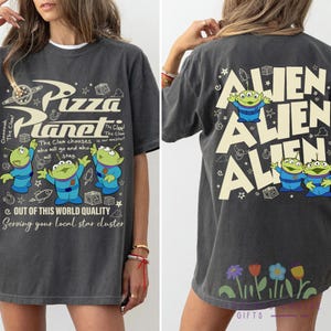 May include: Dark gray t-shirt with a Pizza Planet graphic on the front and Alien characters on the back. The front features the Pizza Planet logo and alien characters. The back has the word "Alien" repeated.