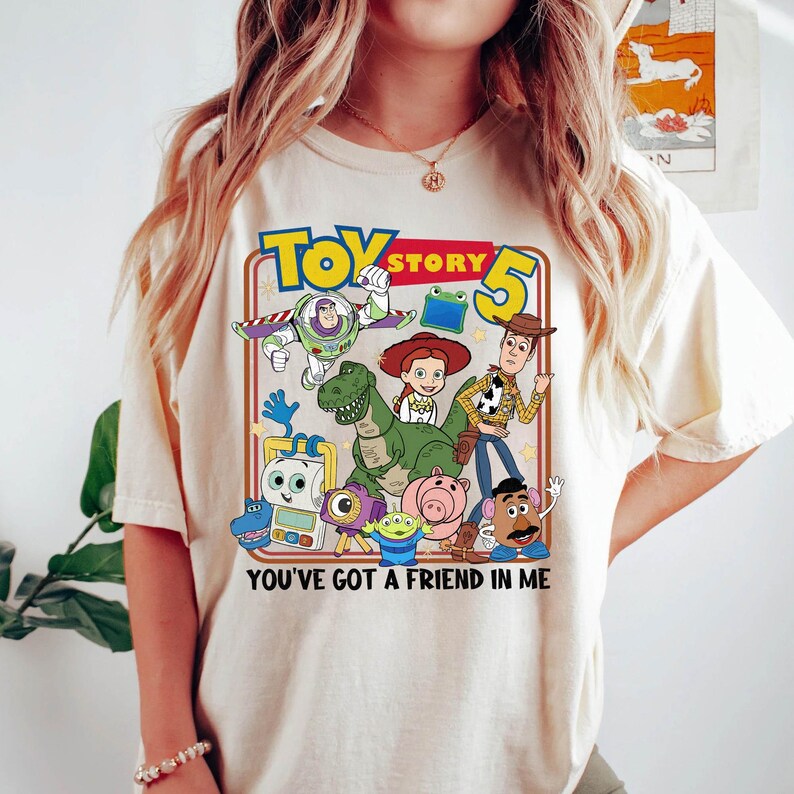 May include: Cream-colored t-shirt featuring a colorful graphic of characters from the animated film "Toy Story 5". The design includes Buzz Lightyear, Woody, Jessie, and other characters, with the text "YOU'VE GOT A FRIEND IN ME".