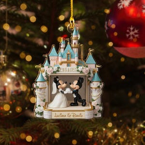 May include: A Christmas ornament featuring a castle design with Mickey and Minnie Mouse figures. The ornament has a light blue and white color scheme, with the word "ENGAGED" displayed. The names "Luciana & David" are also visible. The ornament is hanging from a gold string.