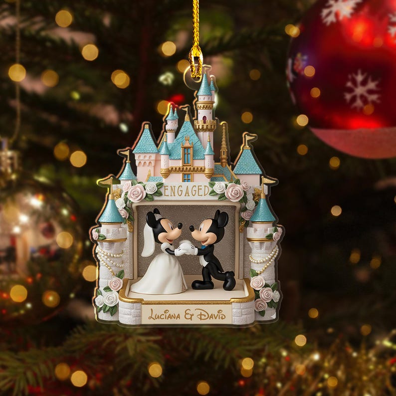 May include: A Christmas ornament featuring a castle design with Mickey and Minnie Mouse figures. The ornament has a light blue and white color scheme, with the word "ENGAGED" displayed. The names "Luciana & David" are also visible. The ornament is hanging from a gold string.