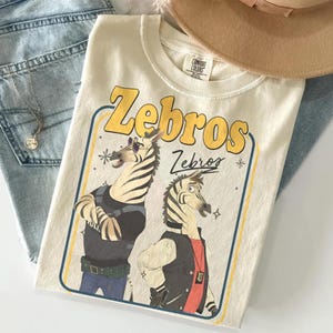 May include: Cream-colored t-shirt featuring the word "Zebros" in yellow, with two cartoon zebras. One zebra wears sunglasses and a black vest, the other a jacket. The shirt is folded on top of blue jeans.