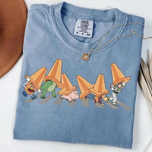 Może przedstawiać: Jasnoniebieska koszulka Comfort Colors z grafiką postaci z Toy Story przechodzących przez przejście dla pieszych, każdy z pomarańczowym pachołkiem drogowym. Złoty naszyjnik jest zarzucony na koszulkę.