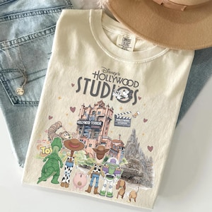 May include: Cream-coloured t-shirt featuring the text "Disney's Hollywood Studios" with a graphic of the Tower of Terror and Toy Story characters. The shirt is paired with blue jeans and a tan hat.