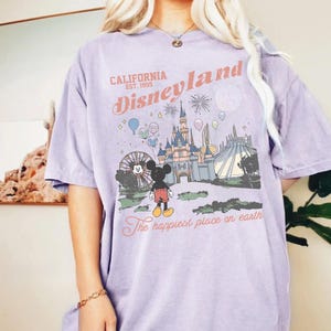 May include: A lilac t-shirt with a Disneyland graphic. The design includes the text "California Disneyland Est. 1995" and an illustration of Mickey Mouse, a castle, and other park elements. The text "The happiest place on earth" is also included.
