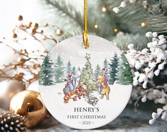 May include: A round Christmas ornament featuring Winnie the Pooh characters gathered around a decorated Christmas tree in a snowy forest scene. The text "HENRY'S FIRST CHRISTMAS ~ 2025 ~" is printed on the ornament.