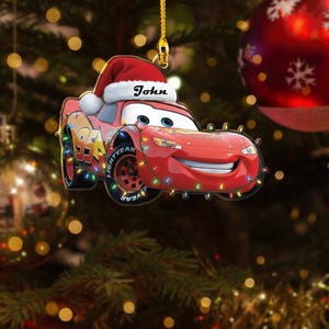 May include: A red Lightning McQueen Christmas ornament with a Santa hat and the name "John" on it. The ornament is decorated with colorful lights and the number 95. The ornament is hanging on a Christmas tree.