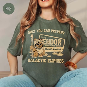 May include: Moss green t-shirt featuring the text "ONLY YOU CAN PREVENT" and a graphic of a bear-like creature holding a spear. The graphic includes the words "Visit ENDOR" and "GALACTIC EMPIRES". The shirt has a relaxed fit and a vintage style.
