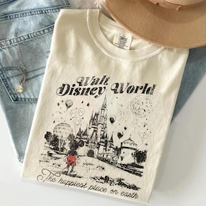 May include: Cream-colored t-shirt with a graphic of Mickey Mouse in front of Cinderella's castle at Walt Disney World. The text reads "Walt Disney World" and "The happiest place on earth". The shirt is folded on top of blue jeans and a tan hat.