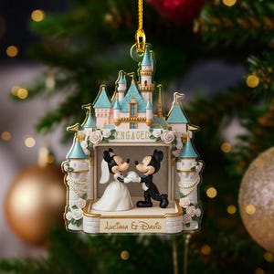 May include: A Disney-themed Christmas ornament featuring Mickey and Minnie Mouse in wedding attire, standing in front of a castle. The ornament is white, pink, and blue, with the word "ENGAGED" and the names "Luciana & David" on it.