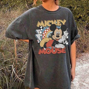 May include: Dark gray t-shirt featuring a vintage-style graphic of Mickey Mouse. The design includes the words "Mickey Mouse" in a retro font, along with a rainbow and stars. The shirt has rolled-up sleeves and a relaxed fit.
