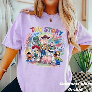 Peut inclure: T-shirt lilas avec l'inscription multicolore "TOY STORY" au-dessus d'un dessin des personnages du film d'animation. Le t-shirt porte &eacute;galement l'inscription "Comfort Colors Orchid" en bas &agrave; droite.