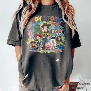 Peut inclure: T-shirt gris fonc&eacute; avec l'inscription "TOY STORY" en lettres color&eacute;es au-dessus d'un dessin des personnages de Toy Story. Le t-shirt a un col rond et des manches courtes. Le texte "Comfort Colors Pepper" est imprim&eacute; en bas &agrave; droite.