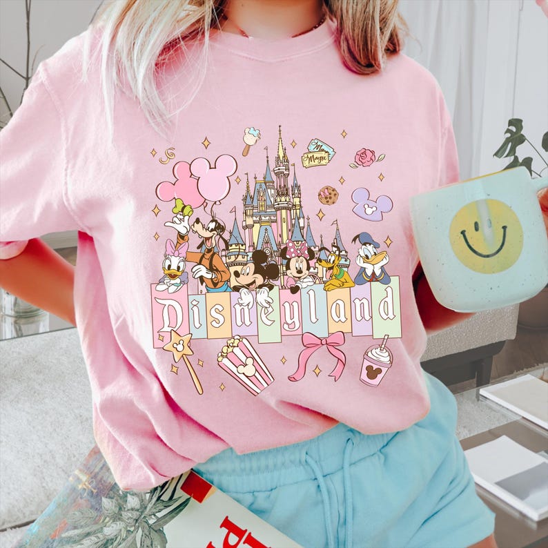 Puede incluir: Camiseta rosa claro con un dise&ntilde;o de Disneyland. El dise&ntilde;o incluye a Mickey Mouse, Minnie Mouse, Donald Duck y otros personajes de Disney, un castillo, globos y la palabra "Disneyland" en colores pastel.