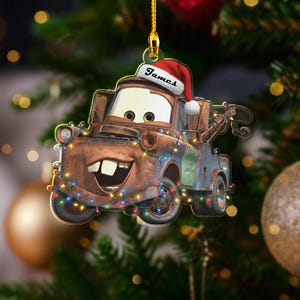 May include: A Christmas tree decoration featuring the character Mater from the film Cars. The ornament is brown and blue, adorned with festive lights, and wears a Santa hat with the name "James". It hangs from a gold string, ready for the festive season.