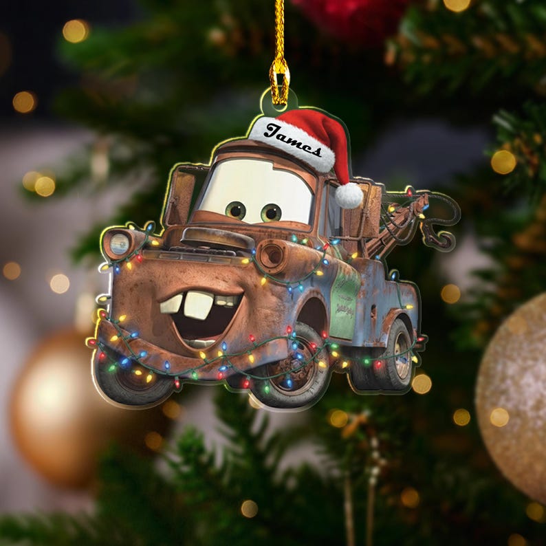 May include: A Christmas tree decoration featuring the character Mater from the film Cars. The ornament is brown and blue, adorned with festive lights, and wears a Santa hat with the name "James". It hangs from a gold string, ready for the festive season.